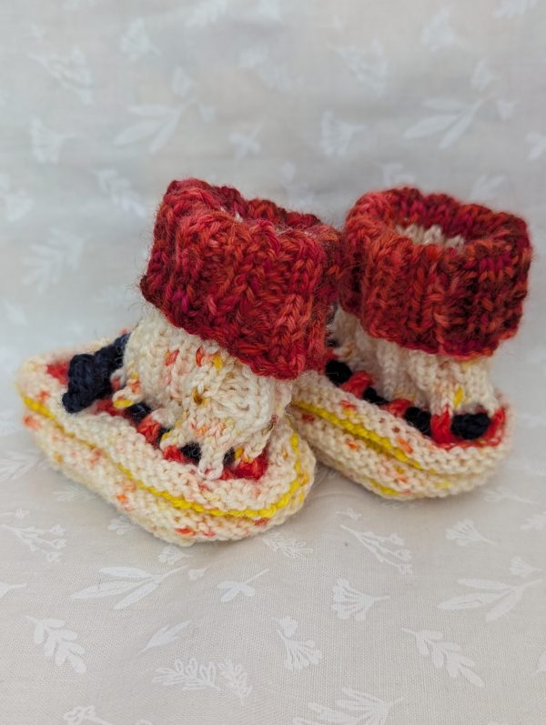 Handknit newborn baby booties in cream, red, and yellow with navy ties and a fiery red cuff, made from soft merino wool.