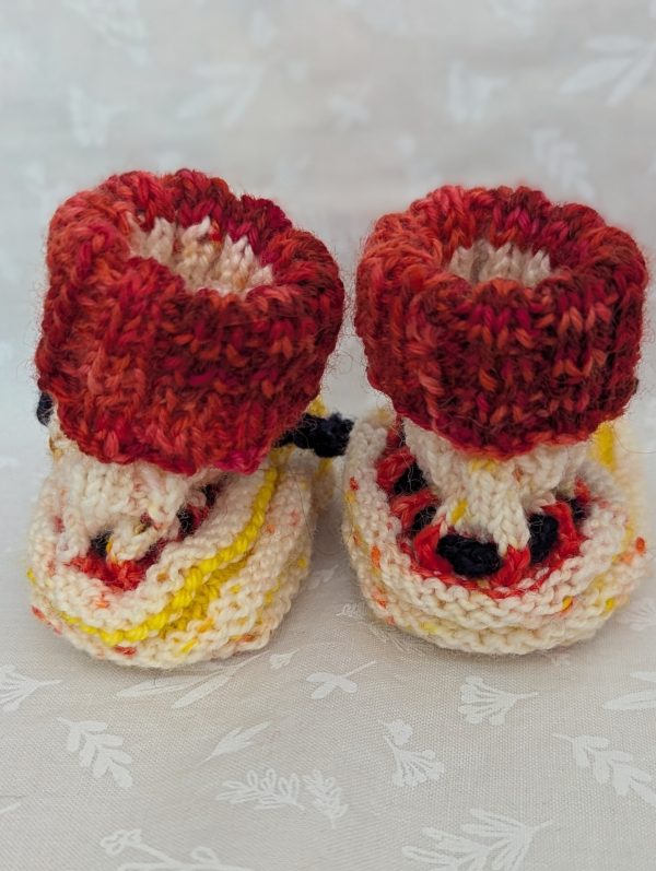Handknit newborn baby booties in cream, red, and yellow with navy ties and a fiery red cuff, made from soft merino wool.