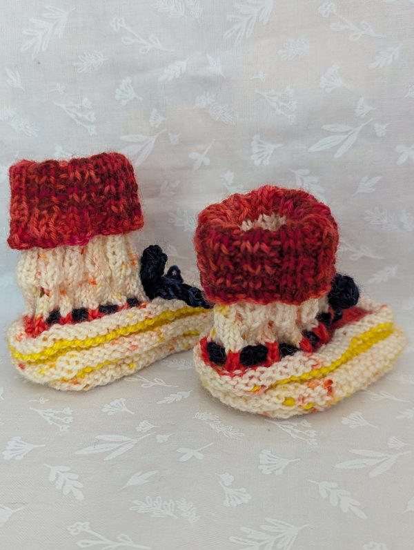 Handknit newborn baby booties in cream, red, and yellow with navy ties and a fiery red cuff, made from soft merino wool.