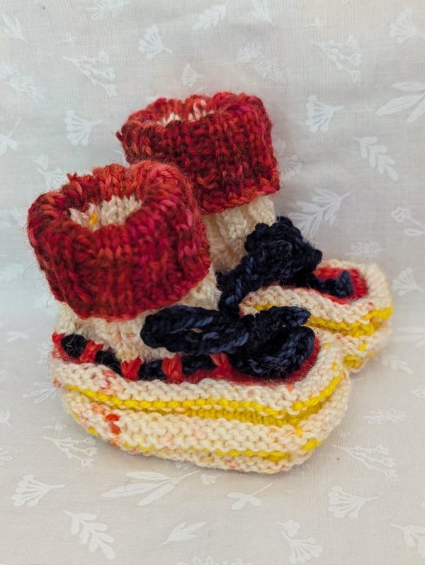 Handknit newborn baby booties in cream, red, and yellow with navy ties and a fiery red cuff, made from soft merino wool.