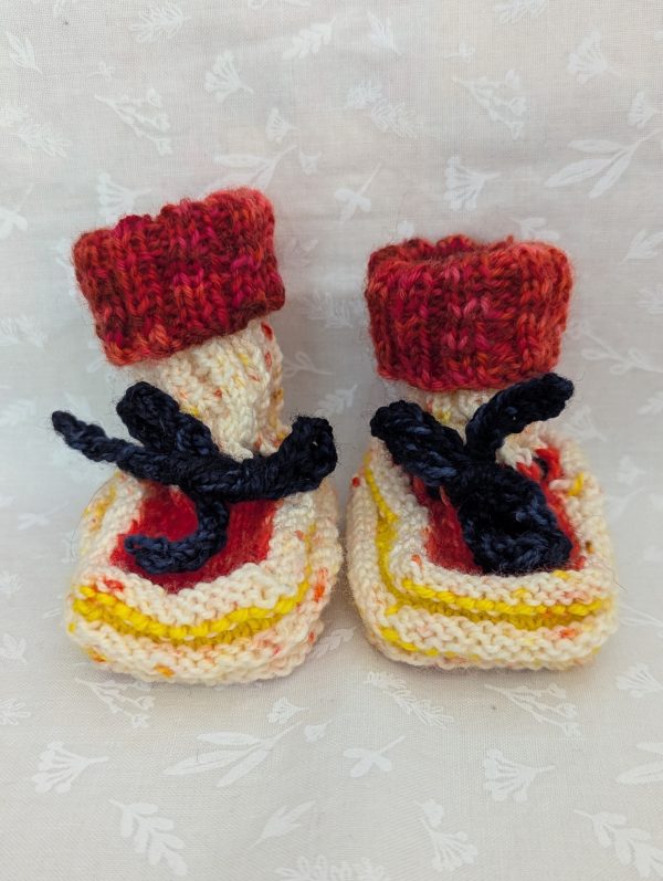 Handknit newborn baby booties in cream, red, and yellow with navy ties and a fiery red cuff, made from soft merino wool.
