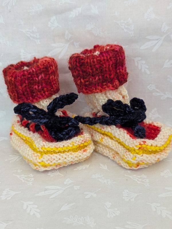 Handknit newborn baby booties in cream, red, and yellow with navy ties and a fiery red cuff, made from soft merino wool.