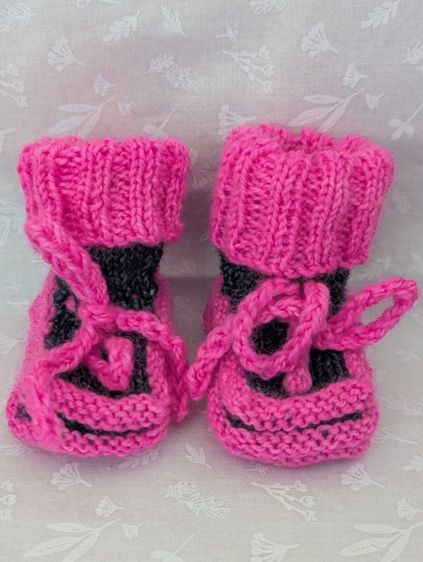 Pink handknit baby booties with blue/black accents, ribbed cuffs, and tied laces, made from soft merino wool for newborns.