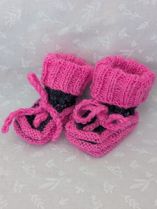 Pink handknit baby booties with blue/black accents, ribbed cuffs, and tied laces, made from soft merino wool for newborns.