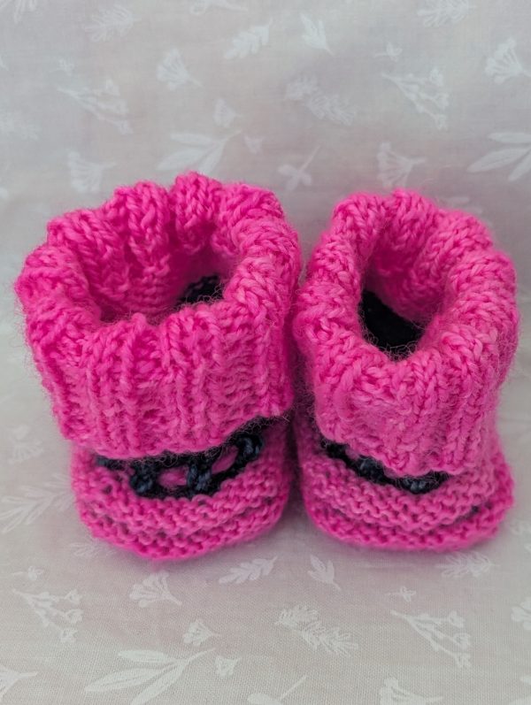 Pink handknit baby booties with blue/black accents, ribbed cuffs, and tied laces, made from soft merino wool for newborns.
