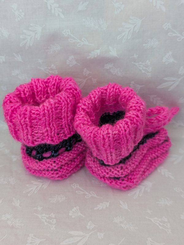 Pink handknit baby booties with blue/black accents, ribbed cuffs, and tied laces, made from soft merino wool for newborns.