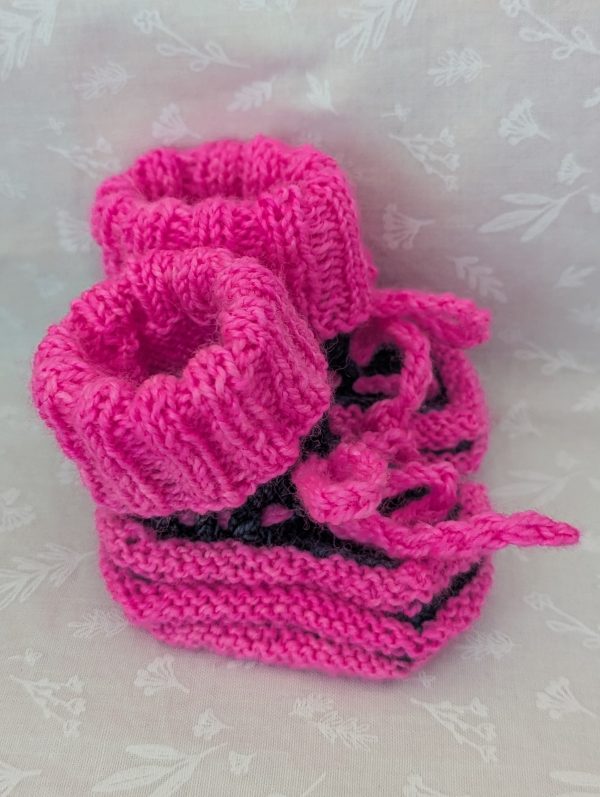 Pink handknit baby booties with blue/black accents, ribbed cuffs, and tied laces, made from soft merino wool for newborns.