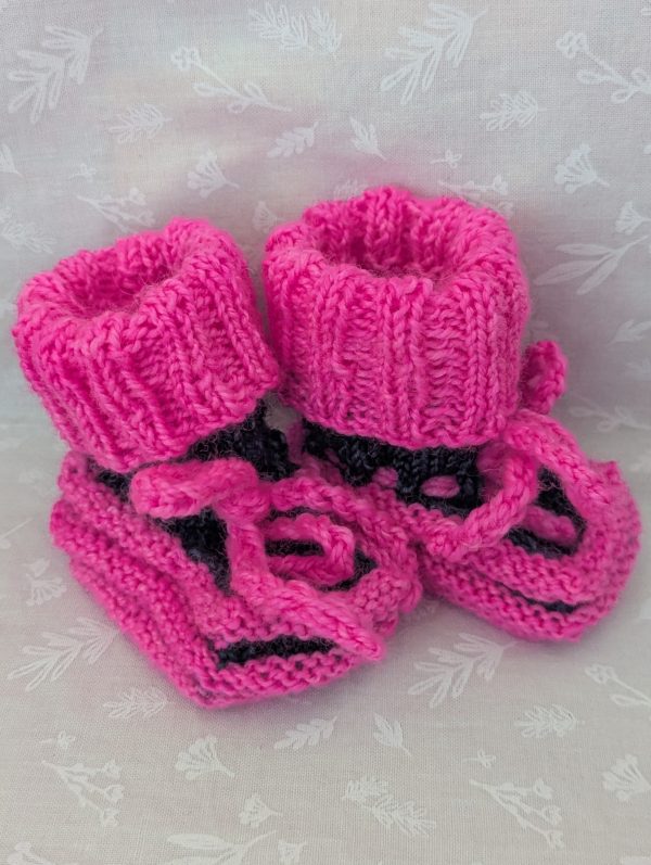 Pink handknit baby booties with blue/black accents, ribbed cuffs, and tied laces, made from soft merino wool for newborns.