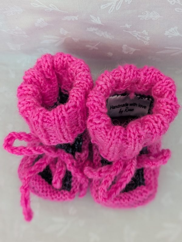 Pink handknit baby booties with blue/black accents, ribbed cuffs, and tied laces, made from soft merino wool for newborns.