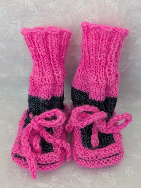 Pink handknit baby booties with blue/black accents, ribbed cuffs, and tied laces, made from soft merino wool for newborns.