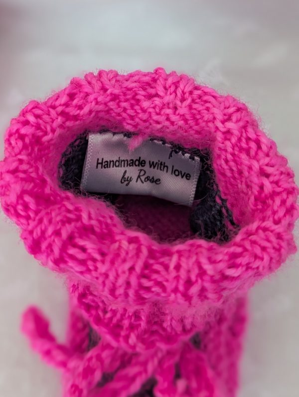 Pink handknit baby booties with blue/black accents, ribbed cuffs, and tied laces, made from soft merino wool for newborns.