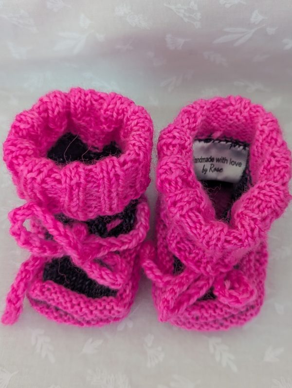 Pink handknit baby booties with blue/black accents, ribbed cuffs, and tied laces, made from soft merino wool for newborns.