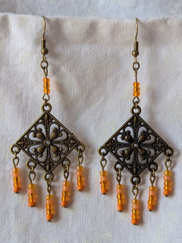 Orange beaded earrings, handcrafted with antiqued bronze square medallions and dangling orange glass beads in five textures, including translucent, frosted, and satin finishes. Hung on bronze ear hooks.