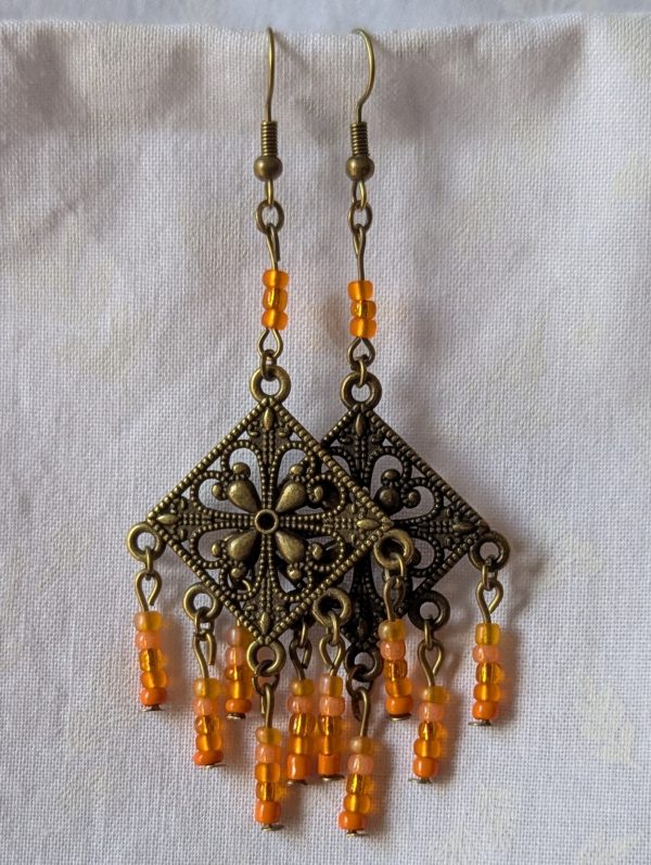 Orange beaded earrings, handcrafted with antiqued bronze square medallions and dangling orange glass beads in five textures, including translucent, frosted, and satin finishes. Hung on bronze ear hooks.