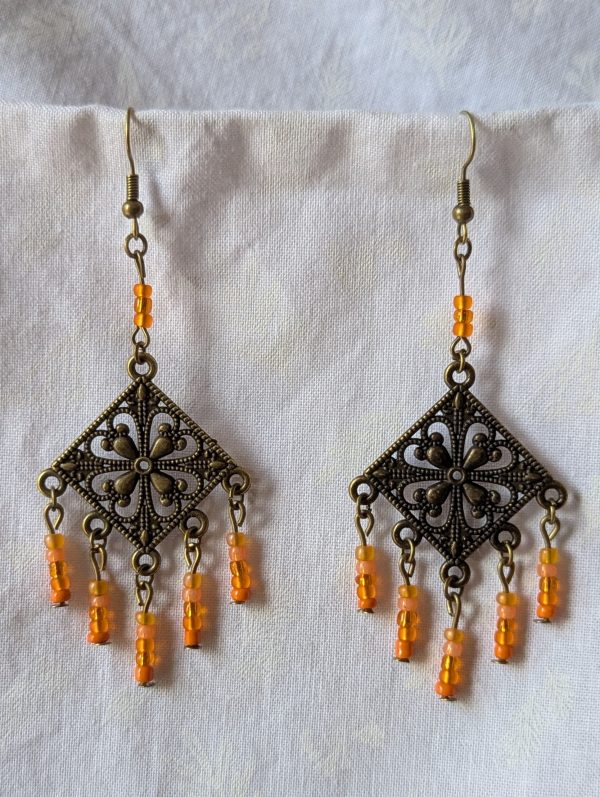 Orange beaded earrings, handcrafted with antiqued bronze square medallions and dangling orange glass beads in five textures, including translucent, frosted, and satin finishes. Hung on bronze ear hooks.