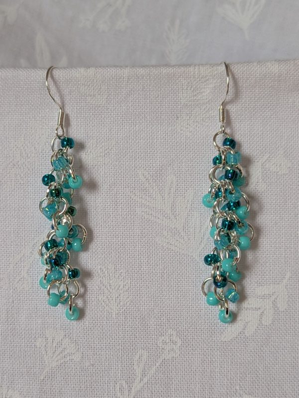 Pair of handmade teal dangle earrings with 27 seafoam, silver-lined teal, and iridescent cyan beads linked by silver rings and sterling silver hooks.