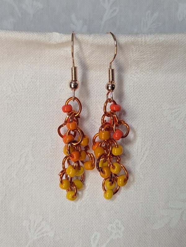 Handmade dangle earrings with 18 orange and yellow glass beads in six warm shades, linked with orange jump rings and rose gold-colored hooks.
