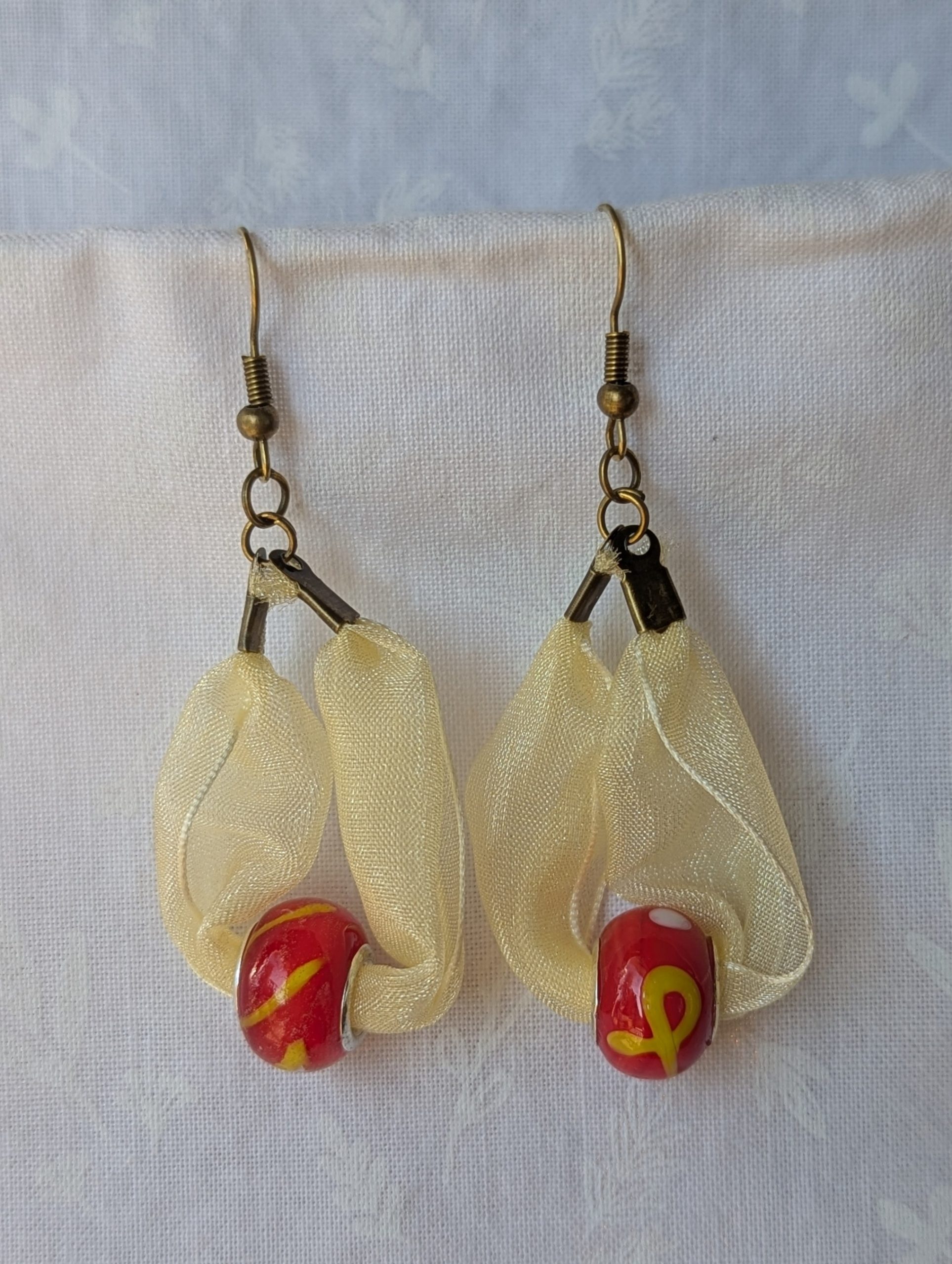 Handmade pale yellow ribbon earrings featuring bright red glass beads with yellow accents and hung from bronze-colored hooks.