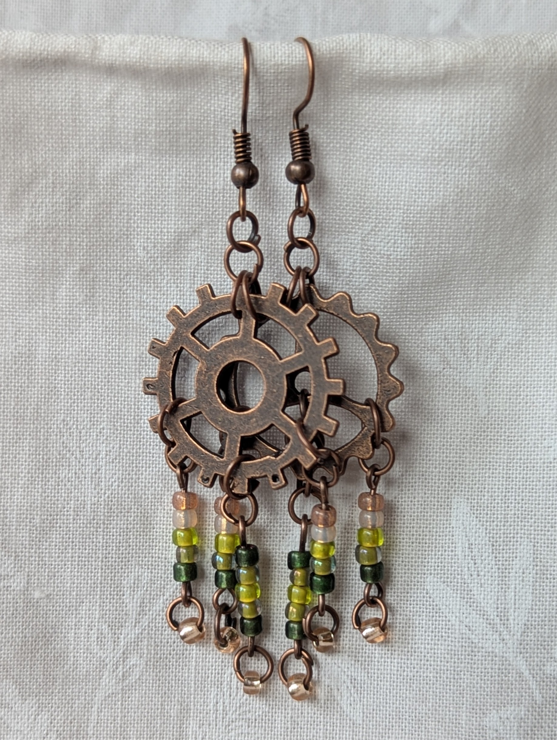 Handmade bronze gear steampunk earrings with green and brown glass beads, asymmetrical steampunk design, and antique bronze hooks.