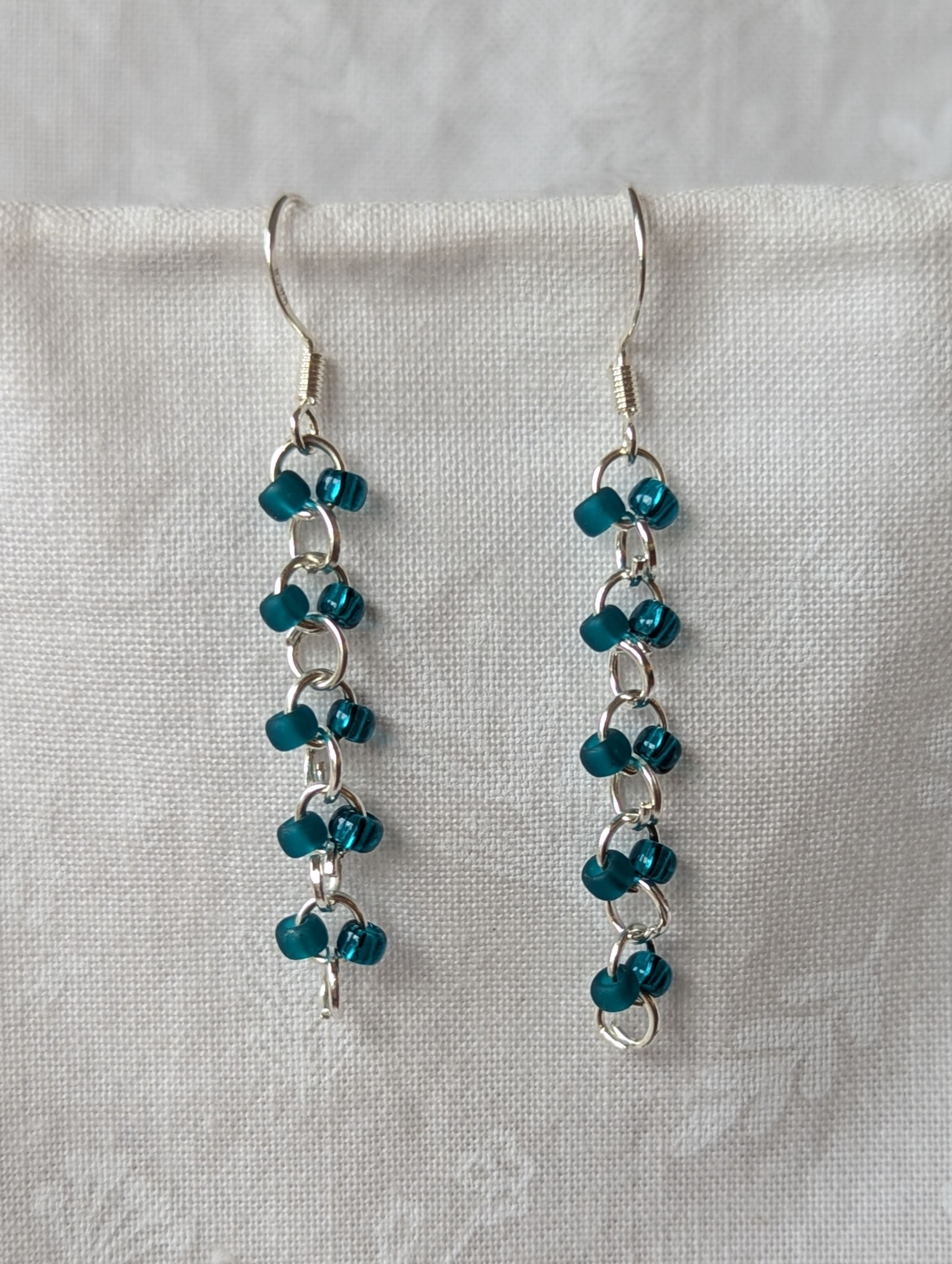 Handmade teal glass earrings featuring frosted and silver-lined teal beads connected with silver jump rings and sterling silver hooks.