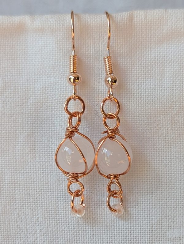 Handmade earrings featuring a milky white glass bead wrapped in copper wire with a white seed bead dangling below, hung from rose-gold colored hooks — Moonlit Copper Glow Earrings.