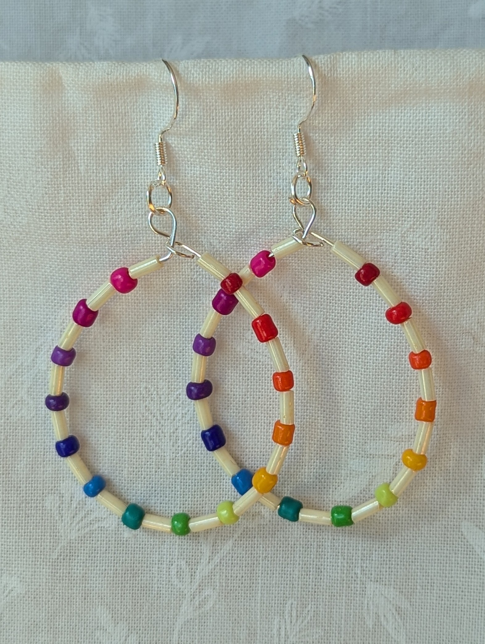 Handmade rainbow and cream accent teardrop shaped loop earrings with alternating colorful glass seed beads and long cream beads on silver-plated wire with sterling silver hooks.