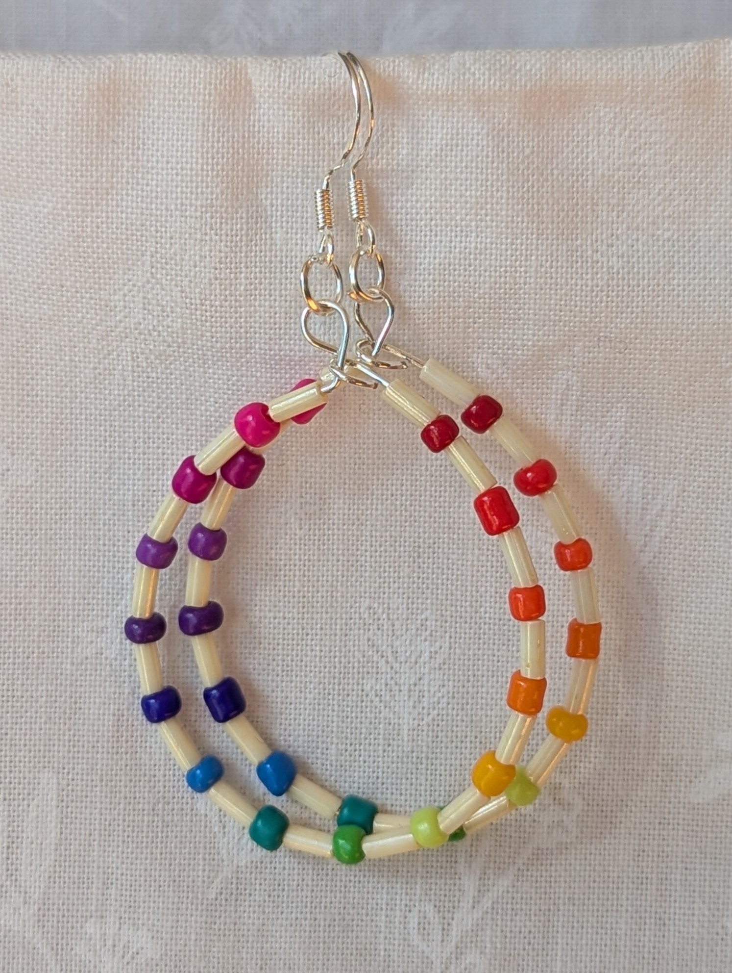 Handmade rainbow and cream accent teardrop shaped loop earrings with alternating colorful glass seed beads and long cream beads on silver-plated wire with sterling silver hooks.
