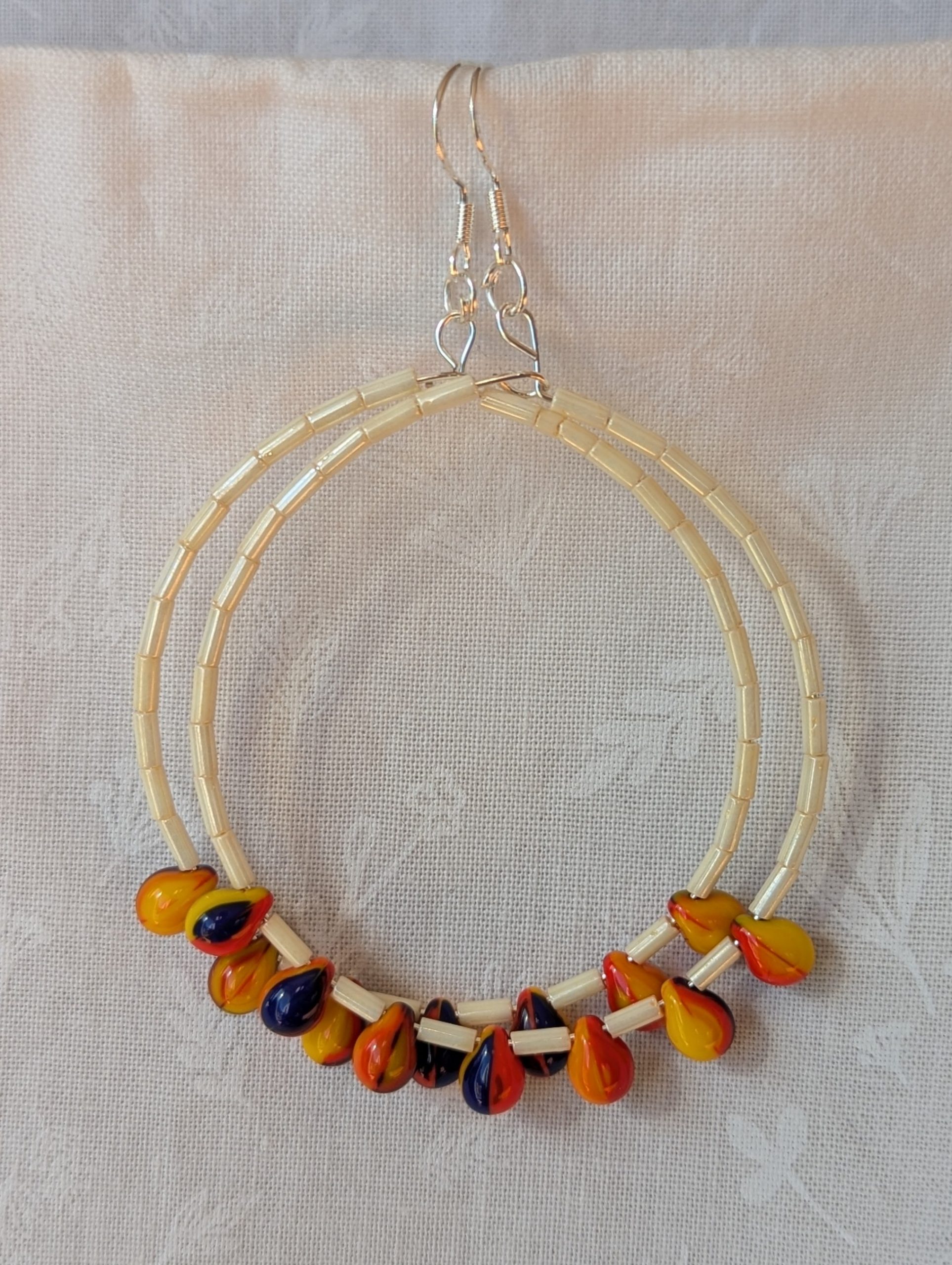 Large cream beaded hoop earrings with multicolor glass teardrop beads in red, yellow, and navy blue on silver-plated wire with sterling silver hooks.