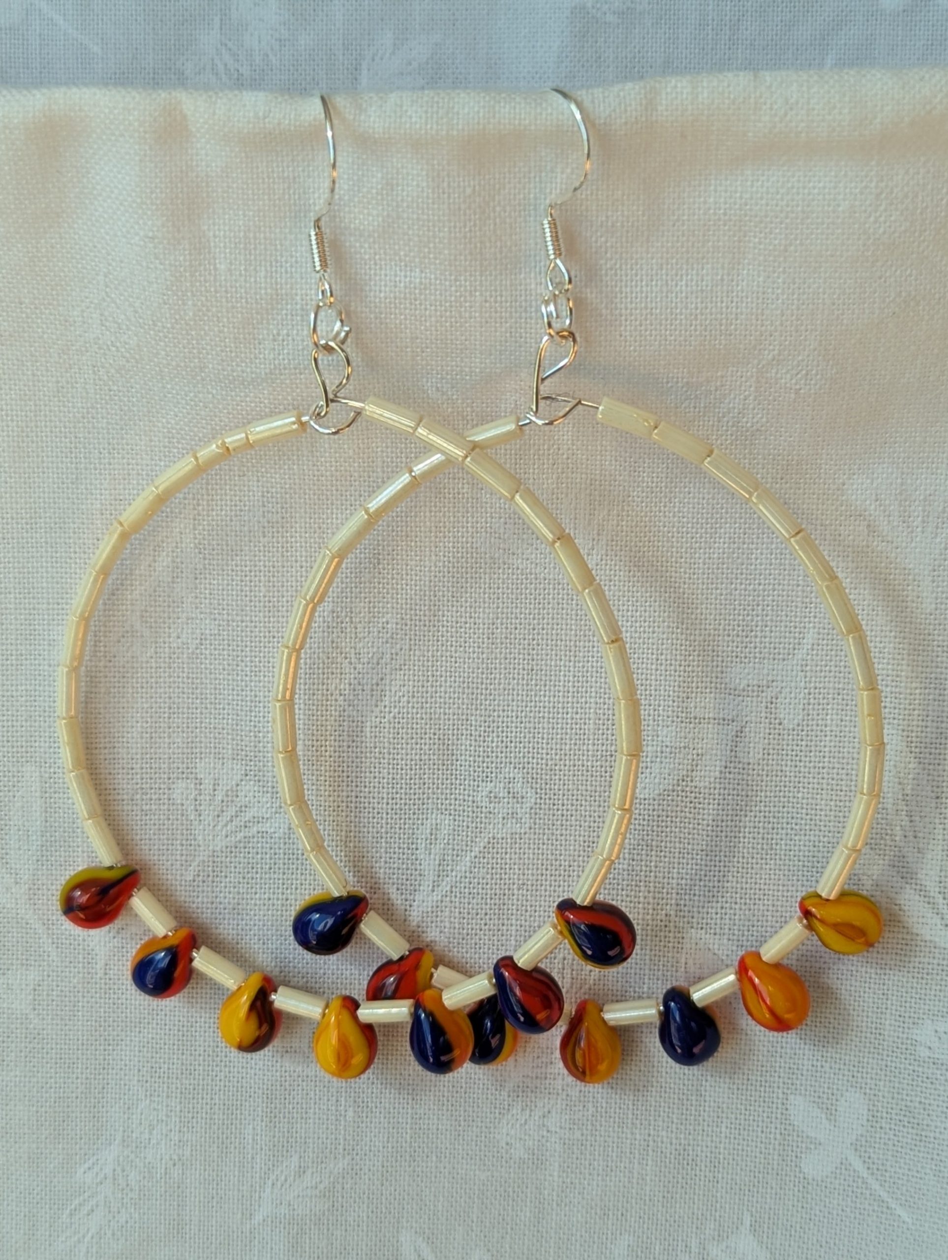 Large cream beaded hoop earrings with multicolor glass teardrop beads in red, yellow, and navy blue on silver-plated wire with sterling silver hooks.